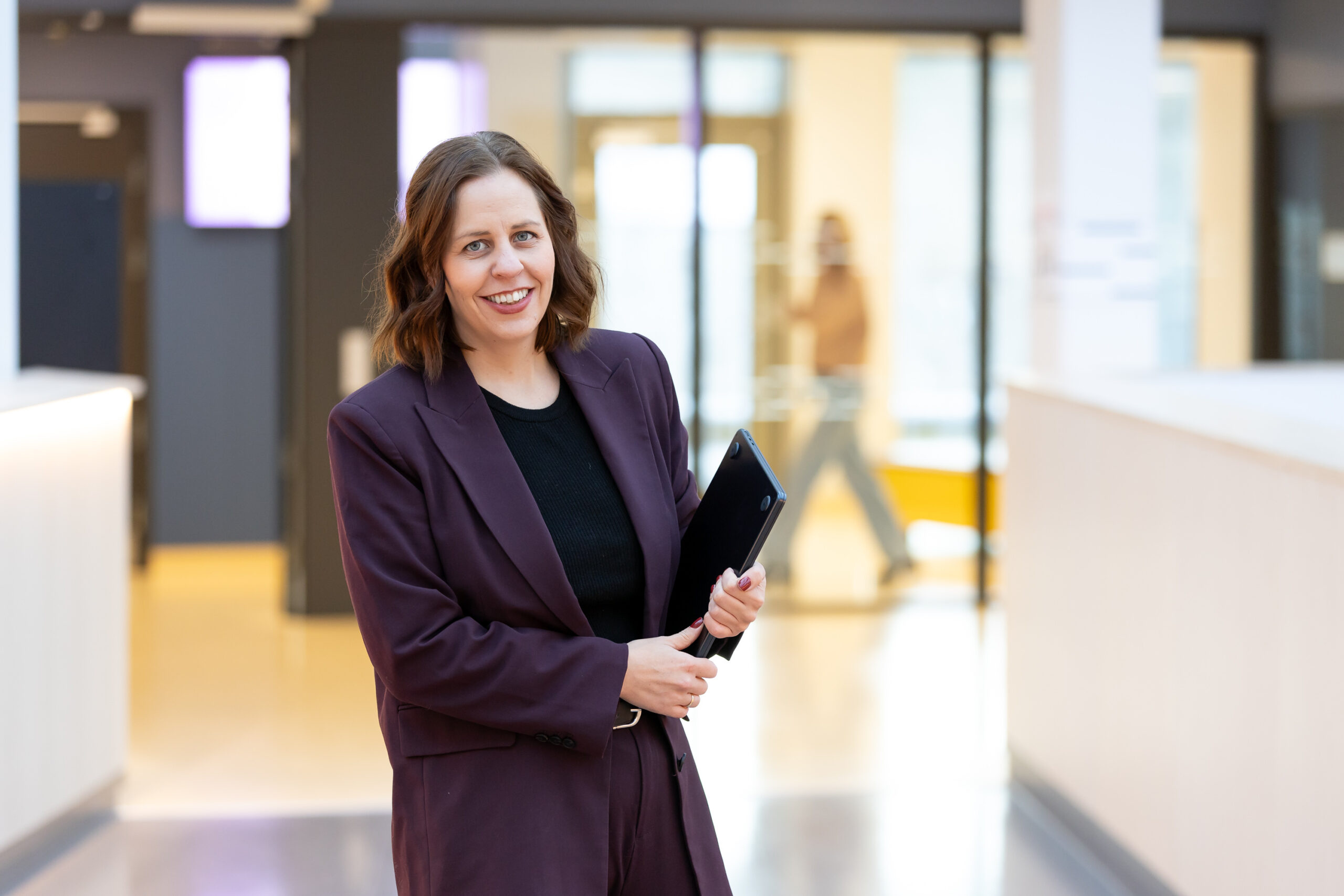 En blazerkledd kvinne i et kontorlandskap smiler og holder en lap top 