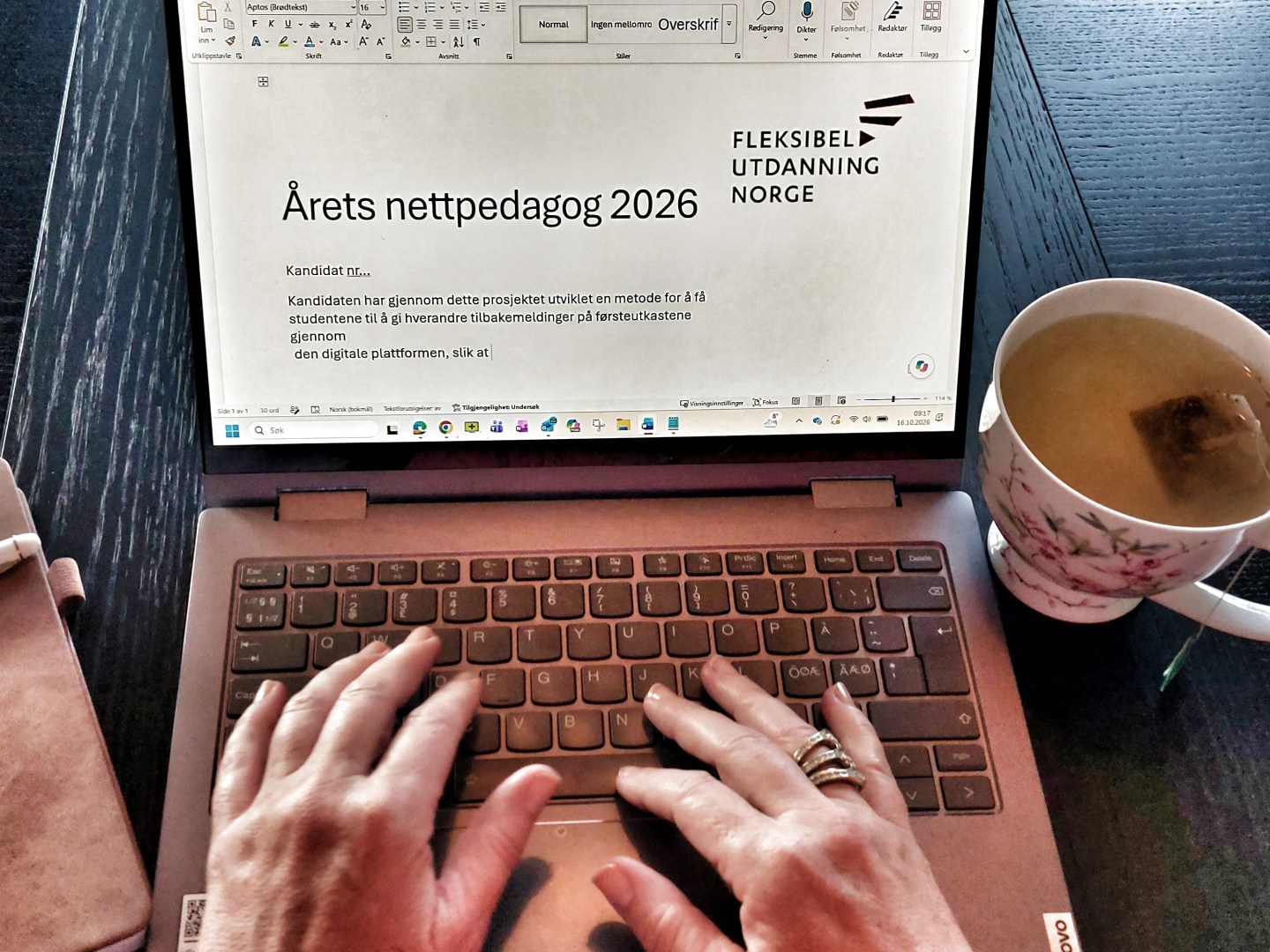 En PC med dokument for Årets nettpedagog. Et par hender skriver på tastaturet. Tekopp og notatbok sees ved siden av.