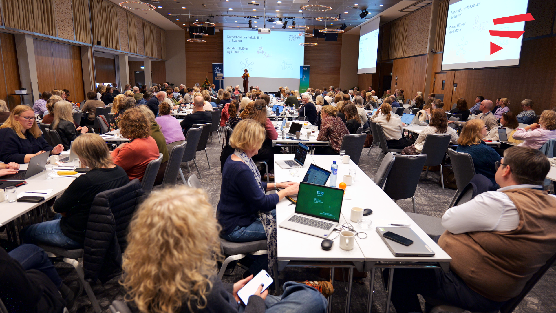 En konferansesal med mange mennesker rundt firkantede bord og en foredragsholder på en scene